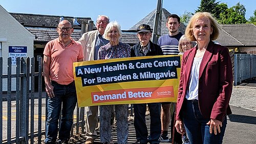 Susan Murray in front of a group of campaigners who are holding up a banner that reads "A new health centre for Bearsden and Milngavie. Demand Better. Scottish Liberal Democrats".