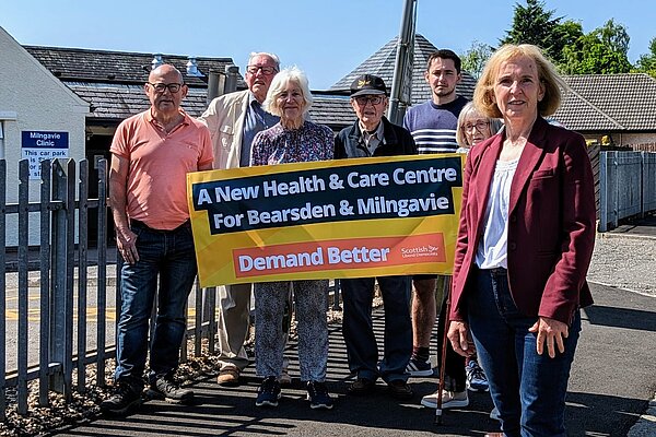 Susan Murray in front of a group of campaigners who are holding up a banner that reads "A new health centre for Bearsden and Milngavie. Demand Better. Scottish Liberal Democrats".