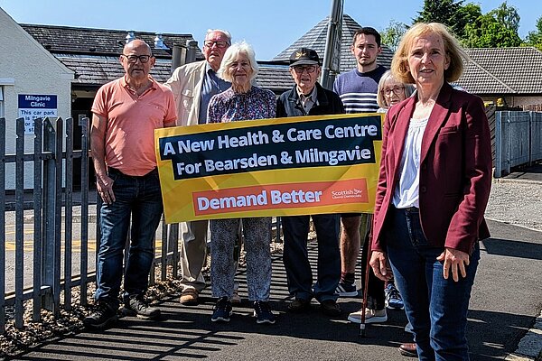 Susan Murray in front of a group of campaigners who are holding up a banner that reads "A new health centre for Bearsden and Milngavie. Demand Better. Scottish Liberal Democrats".