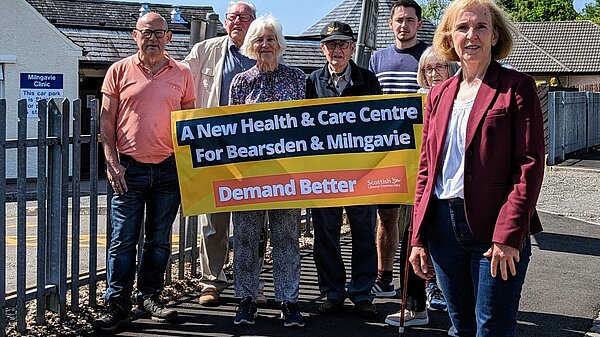 Susan Murray in front of a group of campaigners who are holding up a banner that reads "A new health centre for Bearsden and Milngavie. Demand Better. Scottish Liberal Democrats".