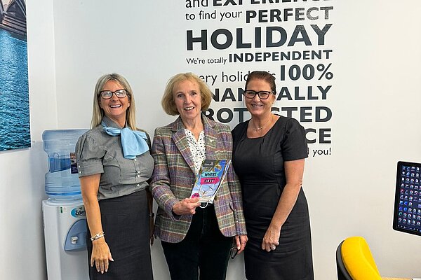 Susan Murray, MP for Mid Dunbartonshire, holding a travel agent leaflet with two ladies smiling at either side.