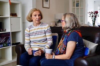 Susan Murray and a resident sitting on a couch chatting. Both of them are holding coffee cups.