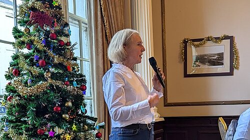 Susan Murray giving a speech, holding a microphone in front of a decorated Christmas tree.